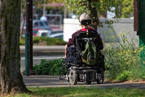 A person using a motorized wheelchair is pictured outside, framed by trees, foliage and in the background, buildings.